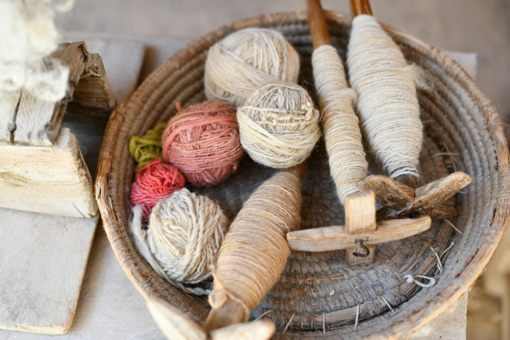 A rustic basket filled with colorful wool yarn balls and wooden spindles for weaving crafts.